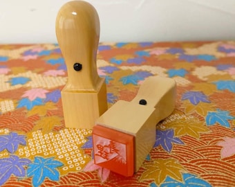May include: Two wooden stamps with black handles. The stamp on the left is plain, while the stamp on the right has a red ink pad with a Japanese character on it.