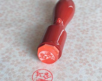 May include: A red wooden stamp with a detailed design on the stamp head. The stamp is angled, showing the red ink impression on a floral patterned paper. The stamp's handle is a deep red color.