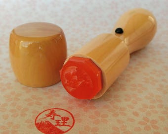 May include: A wooden stamp with a red ink pad and a wooden handle. The stamp has a Japanese character on it. The ink pad is a hexagonal shape. The stamp is on a white background with a floral pattern.