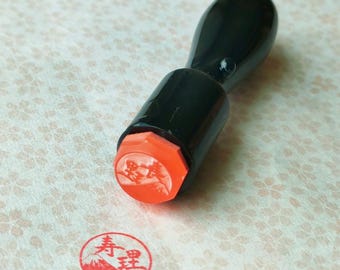 May include: A black wooden stamp with an orange base, used to create a red ink stamp. The stamp has Japanese characters and a mountain design. The stamped image is visible on the paper.