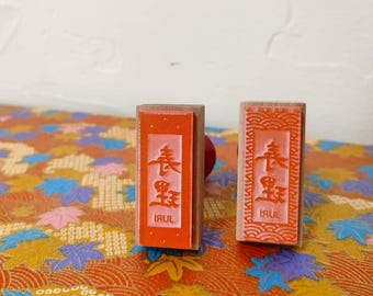 May include: Two red wooden stamps with the Chinese characters for "Spring" and "Festival" in white. The stamps are on a colorful background with a pattern of leaves and flowers.