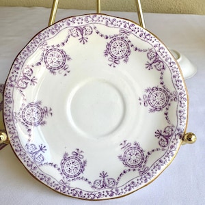 以下が含まれることがあります： 紫色の花柄とメダリオンのデザインが施された白いソーサー。ソーサーは金色の縁とスカラップエッジが付いています。デザインには花の要素と円形のメダリオンが含まれています。ヴィンテージスタイルの作品。