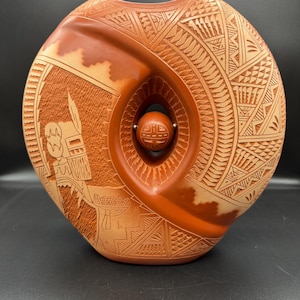 Op de afbeelding: Een handgesneden, terracotta-kleurige keramische vaas met een unieke, afgeronde vorm. De vaas heeft ingewikkelde geometrische en figuratieve ontwerpen, waaronder een centrale opening met een klein, bolvormig element. Het algehele ontwerp is symmetrisch en gedetailleerd.