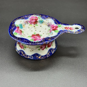 May include: A porcelain tea strainer with a white interior and a blue rim. The strainer is decorated with pink and red roses and gold accents. The handle has a hole for hanging. A decorative item for tea lovers.