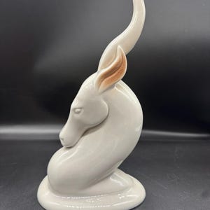 May include: A glossy, off-white ceramic figurine of a gazelle in a resting, curled position. The animal features a single, elongated, curved horn and subtle brown shading on the inner ear, set upon a smooth, oval-shaped base with a reflective finish.