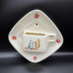 May include: A cream-colored ceramic wall pocket featuring a diamond-shaped backplate with a small hole for hanging. The front displays a molded cup-like container decorated with illustrations of three vases in red, blue, and yellow, plus small red vase motifs.