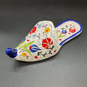 May include: A ceramic slipper-shaped decorative item. The slipper is white with a blue toe and a blue border. It is decorated with red, yellow, and blue floral patterns and green leaves. The slipper is likely a decorative piece.