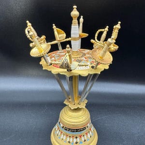 Puede incluir: Un expositor de espadas decorativo de color dorado con múltiples espadas en miniatura. El expositor tiene una base en forma de campana con el texto "TOLEDO-SPAIN" y un diseño de esmalte colorido. Las espadas tienen empuñaduras doradas y hojas plateadas.