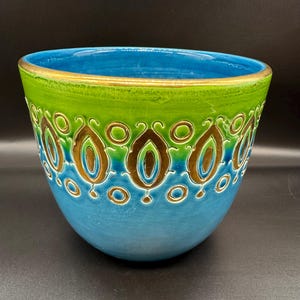 May include: A decorative ceramic bowl with a vibrant colour scheme. The bowl features a turquoise base, a green midsection, and a bright blue interior. The design includes gold and brown teardrop shapes and circles. The rim is trimmed with gold.