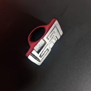 May include: A red and silver ring with a geometric design. The ring has a red band and a silver, rectangular top with a maze-like pattern. The ring is set against a dark background.