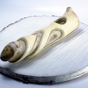 May include: A wooden pipe carved from a branch with a swirling grain pattern. The pipe is light brown and cream colored and has a hole in the side. The pipe is sitting on a white wooden surface.