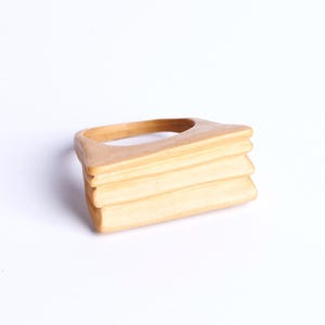 Op de afbeelding: Een houten ring met een gelaagd, rechthoekig ontwerp. De ring is gemaakt van lichtbruin hout en heeft een gladde, gepolijste afwerking.
