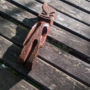 May include: A carved wooden tiki figure with a stylized face and body. The brown sculpture is placed on a weathered wooden surface, with sunlight casting shadows across the piece. The tiki has a distinct, traditional design.