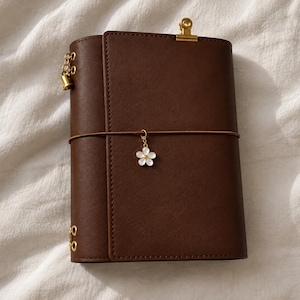 May include: A dark brown leather-like journal cover featuring a gold-toned metal binder clip at the top, a gold-toned bell charm on the side, and a white flower-shaped charm hanging from a thin brown elastic band that wraps around the centre.