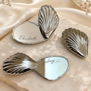 May include: Three silver-toned metal compact mirrors shaped like seashells. Two mirrors are open, revealing reflective surfaces with the engraved names Elizabeth and Abigail in a cursive script. The third mirror is closed, showing a ribbed shell texture.