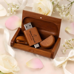 May include: A wooden USB flash drive with the names "Stacey & David" and the date "07-09-2025" engraved on it, along with a wedding ring graphic. The USB drive is inside a wooden box with a matching wooden lid and two wooden pieces. The background includes white flowers and pink petals.