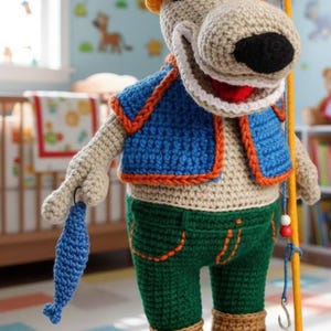 May include: A crocheted dog doll wearing a yellow hat, blue vest with orange trim, and green pants. The dog holds a blue fish and a fishing pole. The doll has a smiling face and is wearing brown boots.
