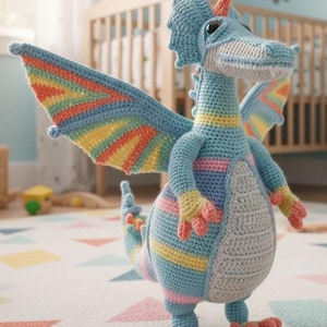 Puede incluir: Un juguete de dragón de ganchillo hecho a mano en tonos de azul, gris, amarillo, naranja y rosa. El dragón tiene grandes alas con rayas arcoíris, un cuello largo y pies pequeños. El juguete está sobre una alfombra estampada.