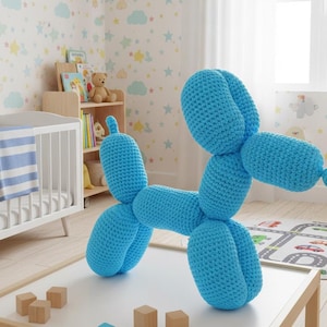 Puede incluir: Un perro globo grande de ganchillo azul claro está sobre una mesa blanca. El juguete tiene cabeza, cuerpo y patas redondeadas, con una pequeña cola. El fondo incluye una cuna, una estantería con libros y una alfombra estampada.