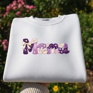 Peut inclure: Sweat-shirt blanc avec le mot "Mama" en lettres patchwork violettes, beiges et blanches. La lettre "M" est ornée d'un nœud, et les lettres ont des motifs floraux et en forme de cœur. Un vêtement décontracté et confortable.