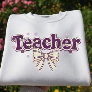Peut inclure: Sweat-shirt blanc avec le mot "Teacher" en violet, orné de motifs floraux et d'un nœud crème. Un vêtement de style col rond, idéal pour un usage quotidien.