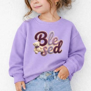 Peut inclure: Un sweat-shirt lilas avec le mot "Blessed" en écriture cursive, accompagné d'un ours en peluche. Les lettres sont marron foncé avec une texture crochetée. L'ours en peluche est marron clair et porte un nœud papillon violet.