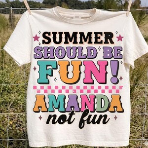Peut inclure: Un t-shirt à manches courtes de couleur crème avec le texte coloré "SUMMER SHOULD BE FUN! AMANDA not fun" dans diverses polices et nuances de violet, bleu sarcelle, orange et rose, agrémenté de petites étoiles, de losanges et d'un motif à damier rose.