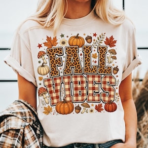 Peut inclure: Un t-shirt couleur crème avec le texte FALL VIBES en grandes lettres majuscules. Le mot FALL a un motif léopard et VIBES a un motif à carreaux rouge et blanc. Le design comprend des citrouilles, des champignons, des feuilles et des glands.