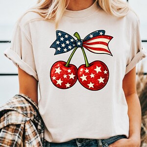 Peut inclure: Un t-shirt beige à manches courtes orné d'un motif représentant deux cerises rouges parsemées d'étoiles blanches. Les cerises sont reliées par des tiges vertes à un nœud aux motifs du drapeau américain, avec du bleu étoilé et des rayures rouges.