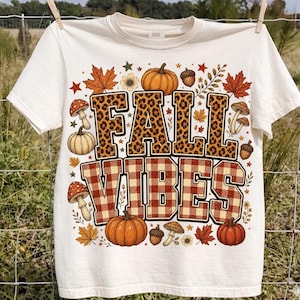 Peut inclure: Un t-shirt à manches courtes de couleur crème avec une impression graphique portant le texte FALL VIBES. Le mot FALL est en motif léopard et VIBES est en motif à carreaux rouge et blanc, entouré de citrouilles, de champignons et de feuilles.