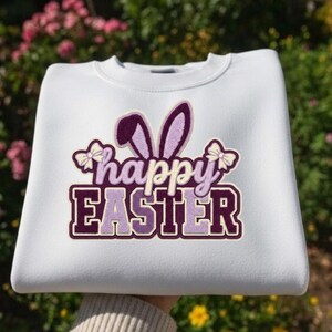 Peut inclure: Sweat-shirt blanc avec un motif "happy EASTER" violet et lavande. Le motif comprend des oreilles de lapin et des nœuds. Le sweat-shirt est en matière douce, parfait pour Pâques.