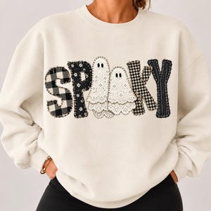 Peut inclure: Un sweat-shirt à col rond couleur crème avec le mot SPOOKY imprimé sur la poitrine. Les lettres sont stylisées avec divers motifs, notamment un carreau vichy noir et blanc, un motif floral noir et deux formes de fantômes en dentelle blanche.