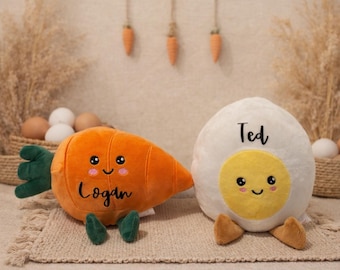 May include: Two plush Easter toys: an orange carrot with the name "Logan" and a white egg with the name "Ted". Both have smiling faces and are on a beige rug. Decorative carrots hang in the background.