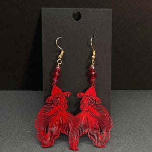 May include: A pair of red fish-shaped earrings. The earrings feature a detailed cutout design of a goldfish with flowing fins. The earrings have gold-colored hooks and are adorned with red beads. The earrings are displayed on a black card.