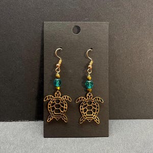 May include: Gold-colored turtle earrings with teal and gold beads. The earrings have a fish hook style closure. The turtle design is open, with a detailed shell and flippers. The earrings are displayed on a black card.