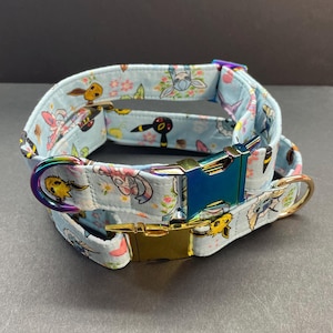 May include: A light blue dog collar with a colorful print of cartoon characters. The collar has a gold and iridescent blue buckle.
