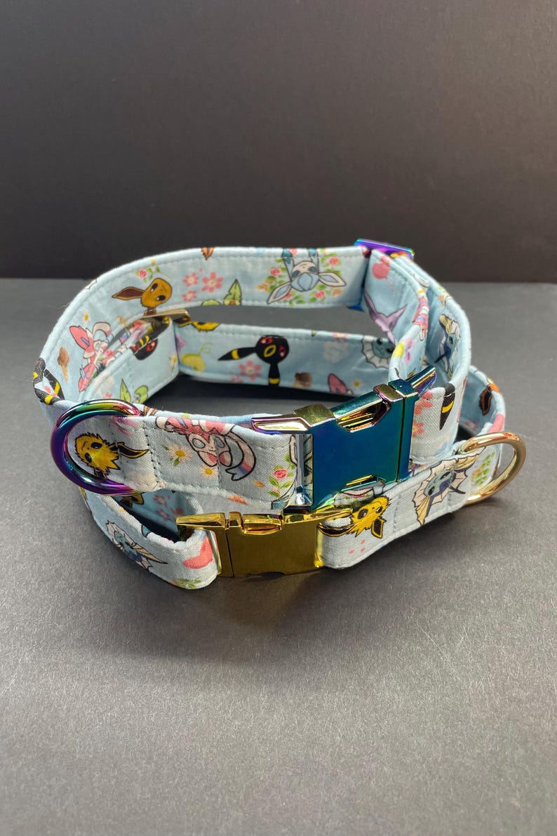 May include: A light blue dog collar with a colorful print of cartoon characters. The collar has a gold and iridescent blue buckle.