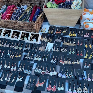May include: A display of handmade earrings, bracelets, and hair accessories on a checkered tablecloth. The earrings feature various designs, including animals, food, and abstract shapes. Wicker baskets and a small orange-patterned bag are also visible.