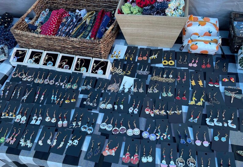 May include: A display of handmade earrings, bracelets, and hair accessories on a checkered tablecloth. The earrings feature various designs, including animals, food, and abstract shapes. Wicker baskets and a small orange-patterned bag are also visible.