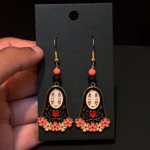 May include: A pair of gold-toned dangle earrings featuring a black figure with a white face, red heart, and pink flower accents. The earrings have gold hooks and are displayed on a black card.