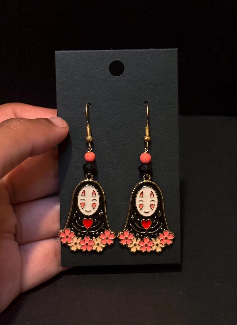 May include: A pair of gold-toned dangle earrings featuring a black figure with a white face, red heart, and pink flower accents. The earrings have gold hooks and are displayed on a black card.