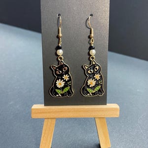 May include: A pair of dangle earrings featuring black cat designs with white daisy accents. The earrings have gold-colored hooks and are adorned with black beads and a white pearl. The earrings are displayed on a black card.