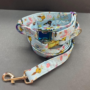 May include: A blue and white dog collar and leash set with a floral pattern and cartoon images of Pokemon characters. The collar has a gold buckle and a rainbow-colored metal D-ring. The leash has a gold hook.