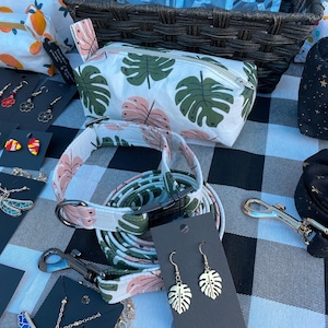 May include: A white and green patterned fabric pouch with a zipper closure. A matching fabric dog collar and leash with a pink trim. A pair of gold earrings with a leaf design. The earrings are on a black card with the text "Monstera Leaf Earrings".