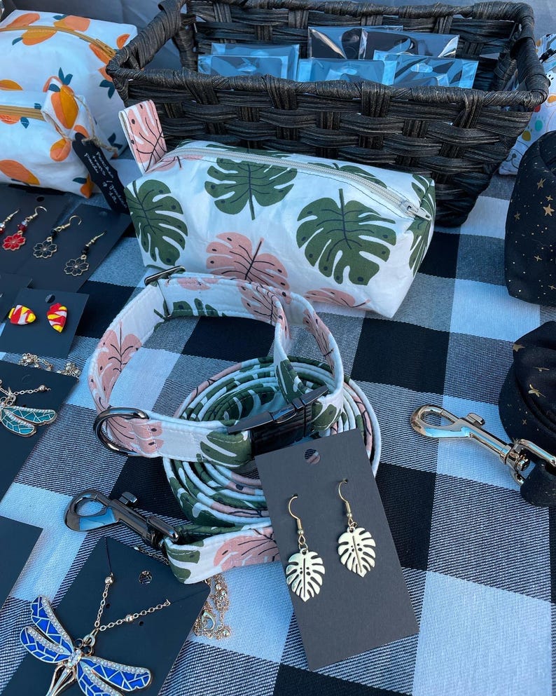 May include: A white and green patterned fabric pouch with a zipper closure. A matching fabric dog collar and leash with a pink trim. A pair of gold earrings with a leaf design. The earrings are on a black card with the text "Monstera Leaf Earrings".
