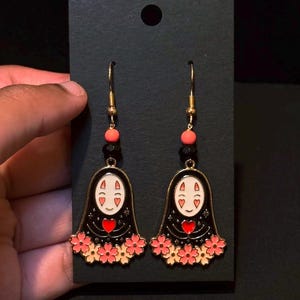 May include: A pair of gold-tone dangle earrings featuring a black design with a white face, red heart, and pink flower accents. The earrings are held against a black background.