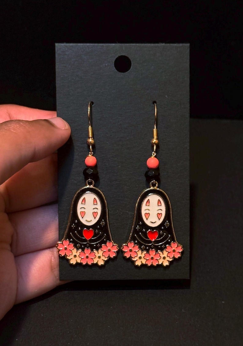 May include: A pair of gold-tone dangle earrings featuring a black design with a white face, red heart, and pink flower accents. The earrings are held against a black background.