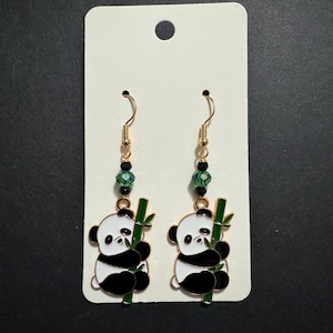 May include: A pair of gold-toned dangle earrings featuring panda bear charms. Each charm depicts a black and white panda holding a green bamboo stalk. The earrings have green beaded accents and are displayed on a white card.