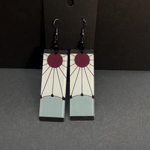 May include: A pair of rectangular earrings with a white background and a red sunburst design. The earrings have a light blue bottom section and black accents. The ear hooks are silver with black beads.