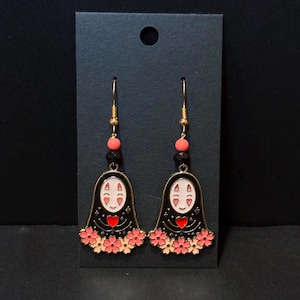 May include: A pair of gold-toned dangle earrings featuring a black figure with a white face and red heart. The earrings have pink flower accents and are suspended from gold hooks with pink and black beads. The earrings are displayed on a black card.